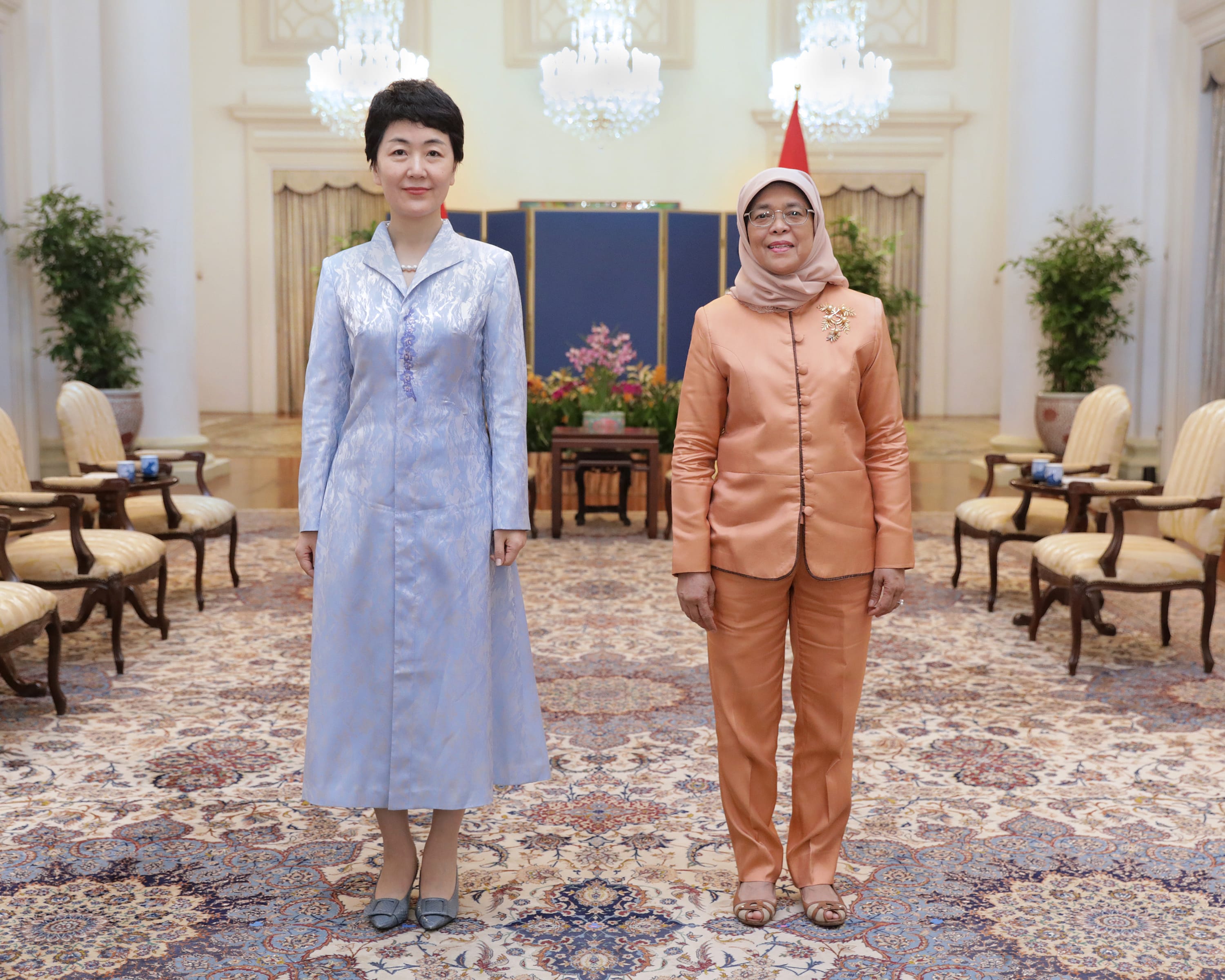 Two women stand in formal attire on an ornate rug in a room with chandeliers.